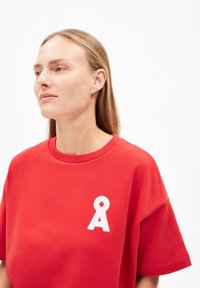 Rotes Baumwoll-T-Shirt mit lockerer Passform, kurzen Ärmeln und einem weißen Logoakzent auf der Brust. Glatte Textur und einfaches Design.