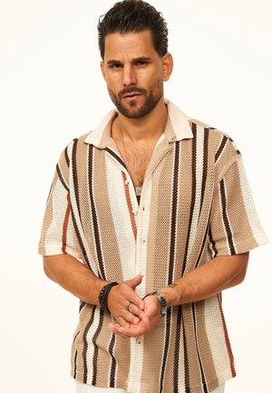 Mann mit kurzen dunklen Haaren und Bart, der ein beige-, braun- und weiß gestreiftes kurzärmeliges Hemd trägt und die Hände hält.
