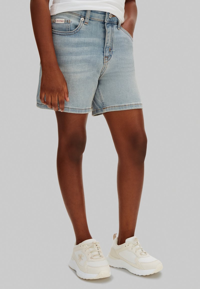 Persona con shorts de mezclilla azul claro, zapatillas blancas y camiseta blanca, de pie con una mano en el bolsillo sobre un fondo gris.