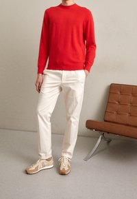 Czerwony sweter z dzianiny, białe dopasowane spodnie, beżowe sneakersy. Prosty design, średnia tekstura i kontrast kolorystyczny. Brązowe krzesło w tle.