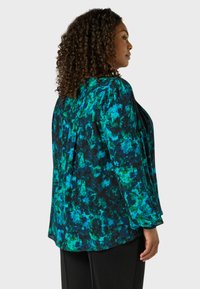 Luftig blus med ett djärvt grönt och blått abstrakt mönster på svart bakgrund. Långa ärmar med subtila rynkor.