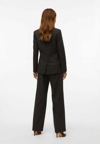 Dunkelgrauer maßgeschneiderter Anzug mit einem taillierten Blazer und weit geschnittenen Hosen. Glattes Material mit einer subtilen Textur und klaren Linien. High Heels.