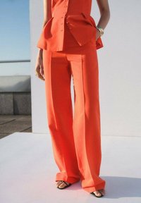 Oranžne hlače s širokimi nogavicami, izdelane iz gladkega materiala. Imajo čist kroj in minimalne šive.