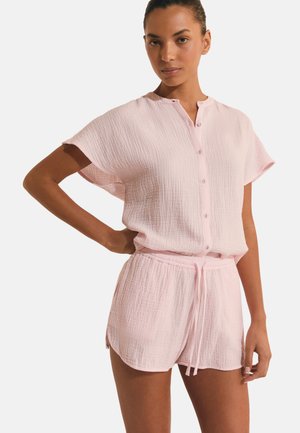 Kobieta w jasno różowej bluzce na krótki rękaw z guzikami i dopasowanych szortach ze ściągaczem wykonanych z teksturowanej tkaniny, stojąca z jedną ręką na biodrze.