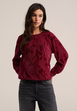 Femme aux longs cheveux foncés portant un chemisier texturé rouge profond à manches longues et un jean gris foncé, debout les mains dans le dos.