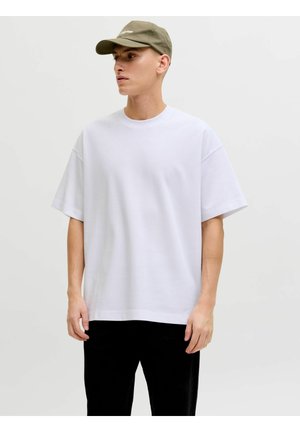 Jeune homme portant un t-shirt blanc oversize, un pantalon noir et une casquette vert olive, debout devant un fond clair uni.