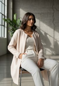 Jasnoróżowy, teksturowany kardigan na białym prążkowanym topie i białych spodniach. Modelka siedzi na krześle, w tle rośliny.