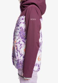 Kvinna i en lila och blommönstrad vinterjacka med huva, står sidledes, "ROXY"-logotypen på ärmen synlig.
