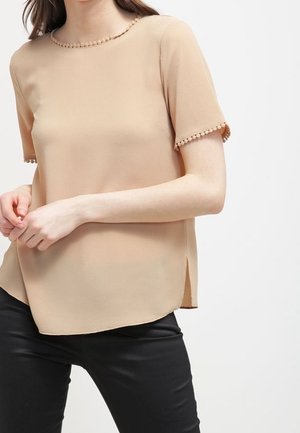 Beige top med korte ærmer i tekstureret stof med pompomkant på ærmer og halsudskæring, båret med sorte bukser.