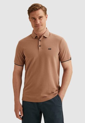 Mann trägt braunes kurzärmeliges Poloshirt mit schwarzen Knöpfen und dunkle Hose, steht mit einer Hand in der Tasche vor einfarbigem Hintergrund.