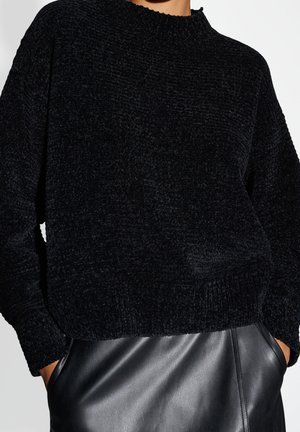 Personne portant un pull noir texturé et un pantalon en cuir noir, les mains dans les poches, sur un fond uni.