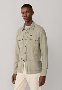 Uomo che indossa una giacca verde chiaro con bottoni e tasche al petto e in vita sopra una maglia grigia, abbinata a pantaloni color crema.