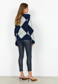 Gestrickter Argyle-Pullover in Marineblau, Grau und Beige mit lockerer Passform; kombiniert mit glänzenden schwarzen Leggings und braunen Mules, die eine elegante Textur zur Schau stellen.