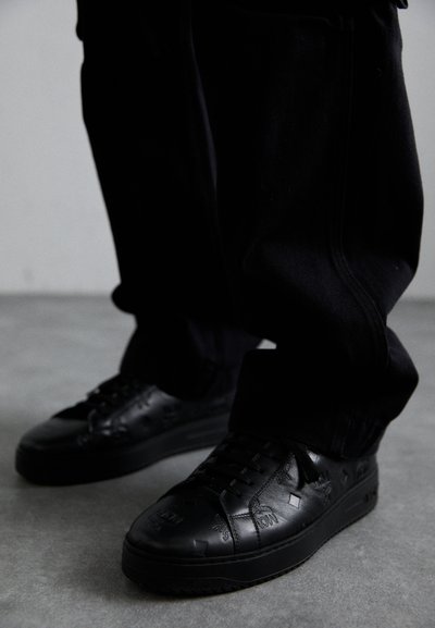 Zapatillas negras de piel con patrones texturizados, que cuentan con una suela de goma y puntera redonda. Los cordones son negros, complementando el diseño oscuro del calzado.