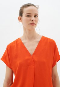 Blusa arancione fluida con maniche corte, scollatura a V e una sottile texture, caratterizzata da un dettaglio di piega frontale.