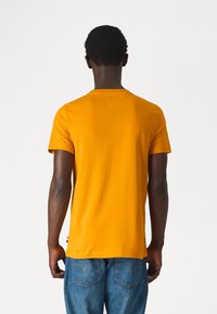 T-shirt arancione in cotone a maniche corte con collo rotondo, dalla texture liscia e con un piccolo logo in accentuazione sull'orlo. Indossato con jeans in denim blu.