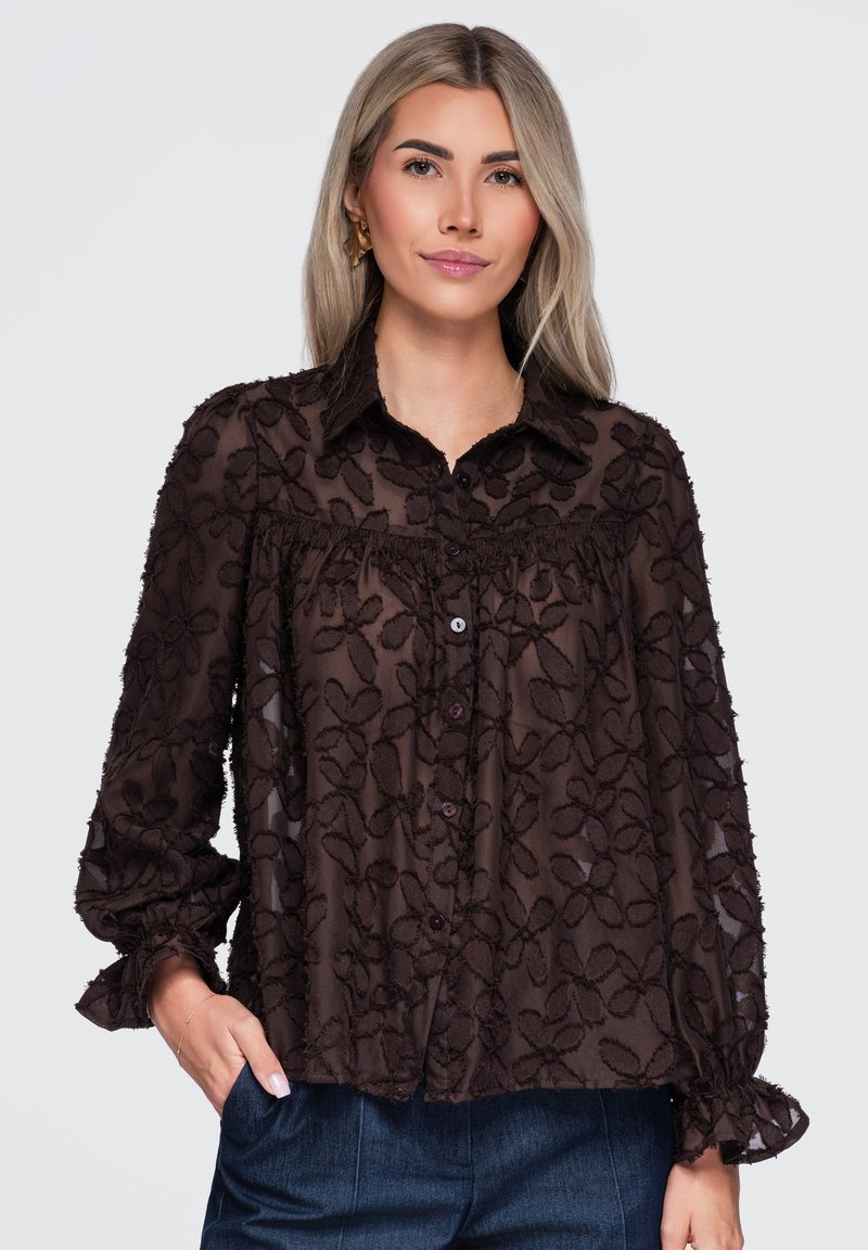 Femme aux longs cheveux blonds portant un chemisier noir transparent brodé de fleurs et un pantalon bleu foncé, une main dans la poche, fond neutre.