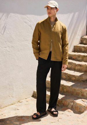 Mann trägt eine hellbraune Jacke mit Reißverschluss, beige Mütze, schwarze Hose und schwarze Sandalen, steht neben Steinstufen an weißer Wand.