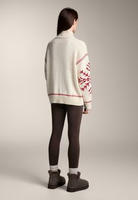 Cremefarbener, gestrickter Pullover mit rotem Schneeflockenmuster, hohem Kragen und geripptem Saum. Kombiniert mit braunen Leggings und grauen Stiefeln mit Pelzbesatz.