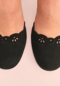 Sorte ruskindssko med bølget kant og laserudskårne blomsterdesigns. Skoene har en glat tekstur og rund tåform.