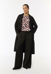 Schwarzer übergroßer Mantel, Bluse mit rosa und schwarzem Zebra-Muster, hoch geschnittene schwarze Hose mit gefaltetem Vorderteil, glatter Stofftextur.