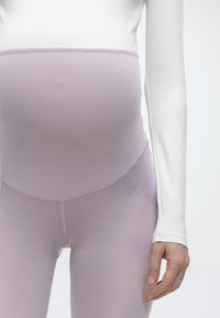 Έγκυος που φοράει λιλά παντελόνι εγκυμοσύνης και λευκό μακρυμάνικο τοπ, εμφανίζεται από τη μέση μέχρι τους γοφούς.