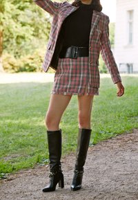 Vaaleanpunainen ja musta ruudullinen bleiseri, joka on paritettu samanlaisen mini-hameen kanssa, lisättynä korkeilla mustilla nahkasaappailla, joissa on ketjukoristeet. Pintakuvioitu kangas ja muotoiltu istuvuus.