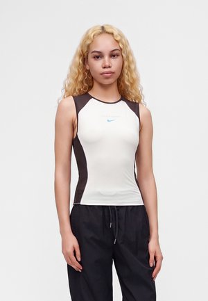 Ung kvinde med langt krøllet blondt hår iført en ærmeløs hvid og sort Nike-top og sorte bukser, stående mod en ensfarvet baggrund.