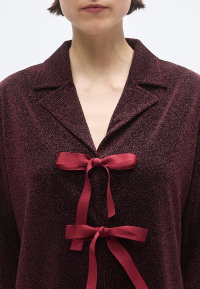 Mørkerød bluse med blank finish, der har en krave og to bånd, som bindes foran. Glat stof med en diskret glans.