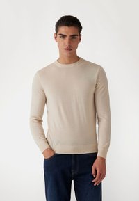 Beiger, gestrickter Pullover mit langen Ärmeln und rundem Ausschnitt, feiner Textur und gerippten Bündchen, kombiniert mit dunkelblauen Jeans.