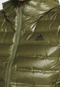 Błyszcząca oliwkowa zielona kurtka ocieplana z zamkiem błyskawicznym z przodu i czarnym logo Adidas po lewej stronie klatki piersiowej.