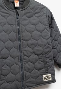 Gewatteerd babyjasje in donkergrijs, met een rits aan de voorkant, geribde kraag, zijzak en een aangehecht label met tekst en een bergafbeelding.