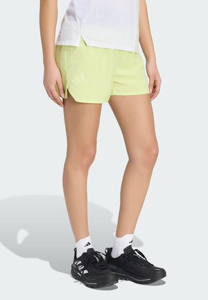 Dolna część ciała osoby noszącej jasnożółte sportowe szorty, białe skarpety Adidas i czarne buty do biegania na jednolitym tle.