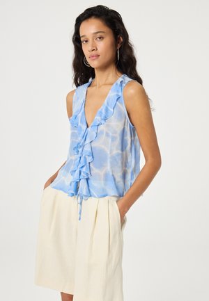 Vrouw draagt een mouwloze blauwe blouse met ruches en tie-dye patroon en hoge taille crème shorts, staat met handen in de zakken tegen een witte achtergrond.