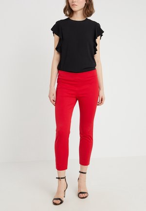 Femme portant un haut noir à manches à volants, un pantalon rouge vif longueur cheville et des sandales noires à talons hauts avec bride à la cheville, debout sur un sol blanc.