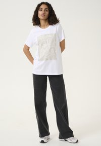 T-shirt en coton blanc avec un grand design graphique beige clair sur le devant et un jean noir à jambe large. Modèle portant des baskets blanches.