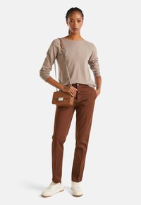 Kobieta w beżowym swetrze, brązowych spodniach, białych sneakersach, trzymająca małą brązową torebkę na ramię z srebrnym łańcuszkiem.