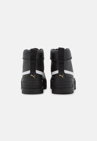 Svartvita hög-top sneakers med guld Puma-logotyp på hälen, sedd från baksidan, mot en plain ljusgrå bakgrund.