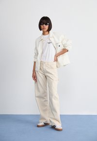 Beige wide-leg jeans, white t-shirt with black logo, off-white jacket, and black sunglasses; casual footwear features a white body and brown sole.