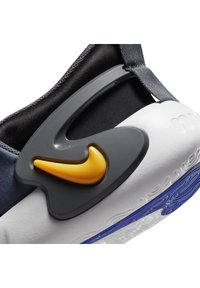 Gros plan sur le talon d'une basket avec un support en plastique gris, un logo Nike jaune, une semelle intermédiaire blanche et une semelle extérieure bleue, montrant un design de semelle texturé.