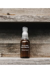 Brązowa szklana butelka z przezroczystą pompką u góry; oznaczona napisem "SUSANNE KAUFMANN Serum Odżywcze" w białym kolorze na ciemnym tle.