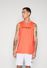 Orange atletisk tanktop med ordet "HYROX" i sort, parret med hvide shorts. Stoffet ser let og glat ud.
