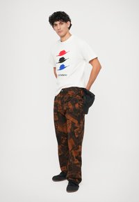 Weißes T-Shirt mit grafischem Druck in Rot, Schwarz und Blau; braune gemusterte Hose mit Zeitungsdesign; schwarze Schuhe; lässiger Schnitt.