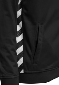 Svart sportjacka med sidostripningar som har vita och svarta diagonala ränder. Inkluderar en framficka och ribbad midja.