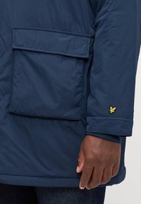Giacca isolante blu navy con una grande tasca a patta sul fronte, polsini a coste e un piccolo emblema dorato sulla manica.
