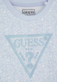 Svetlomodré bavlnené tričko s textúrovaným, irizujúcim trojuholníkovým logom s nápisom "GUESS U.S.A." a pozadím s kvetinovým vzorom.