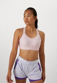 Sujeción deportiva de color rosa claro con escote redondo y tirantes anchos, combinada con pantalones cortos deportivos de color púrpura claro hasta el medio muslo, que cuentan con una cinturilla blanca.