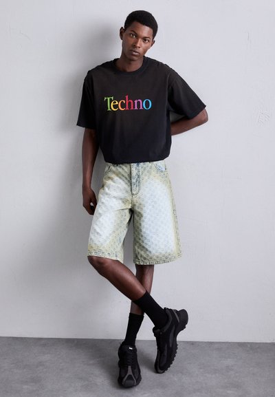 T-shirt noire oversize avec "Techno" en lettres arc-en-ciel ; shorts en denim à motifs bleu clair ; baskets et chaussettes noires. Fond uni.