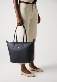 Marineblå totebag med doble håndtak, glidelåslukking, gulllogo, glatt tekstur, kombinert med lyse bukser og ballettsko.