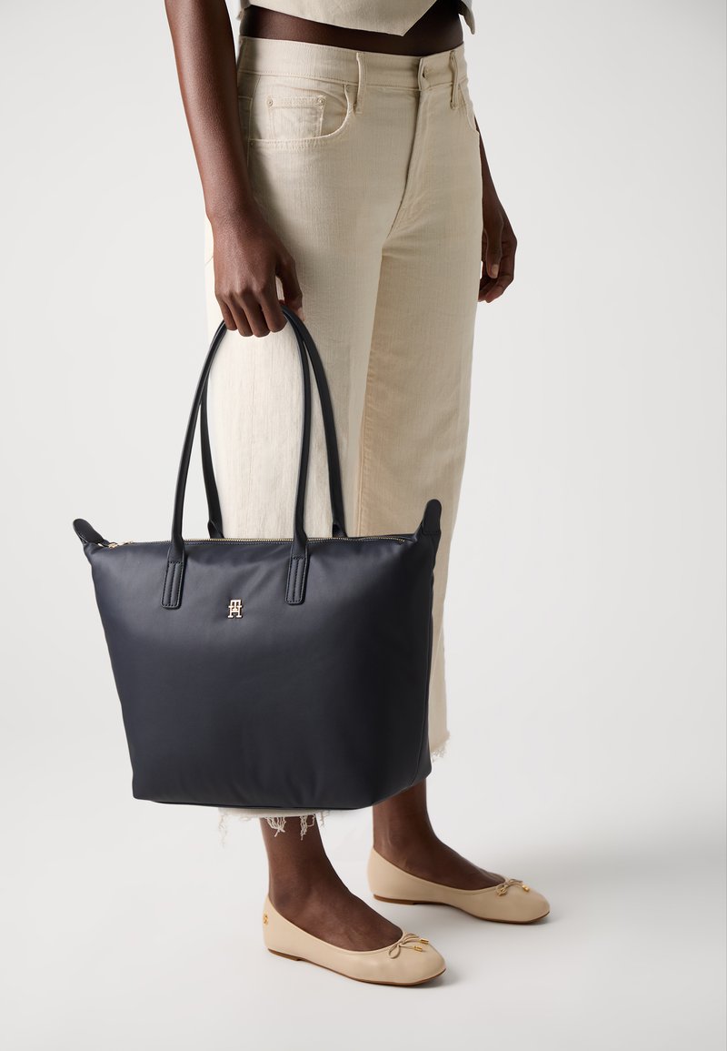 Marineblå totebag med doble håndtak, glidelåslukking, gulllogo, glatt tekstur, kombinert med lyse bukser og ballettsko.
